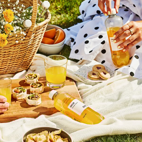 Escena de picnic con bebida de naranja ISKUB y bocadillos en una manta. La persona sostiene una botella de la bebida.