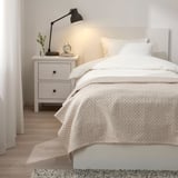Dormitorio minimalista con cama blanca, colcha JAKOBSLILJA, mesita de noche con lámpara y reloj. Decoración ligera.