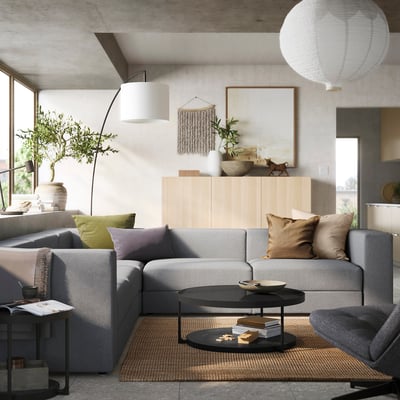 Sala de estar moderna con sofá modular JÄTTEBO gris, consola de madera, planta en maceta, luces colgantes y cojines de colores.