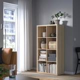 Estantería de madera KALLAX con libros, plantas, decoración. Silla de cuero marrón, ventana con cortinas.