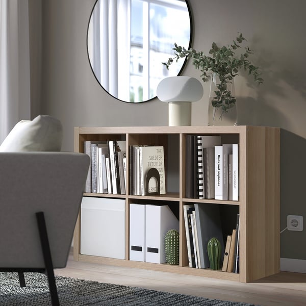 Una estantería de roble claro KALLAX con varios artículos. Es una unidad de tamaño mediano junto a una silla gris, con libros, una lámpara blanca y un jarrón con vegetación.