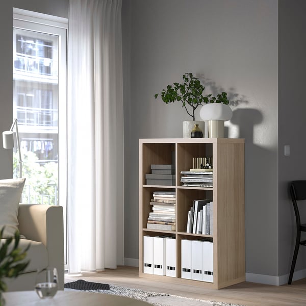 Estantes KALLAX, de madera clara, cerca de la ventana, que sostienen libros y decoración. Líneas altas, sencillas y limpias.
