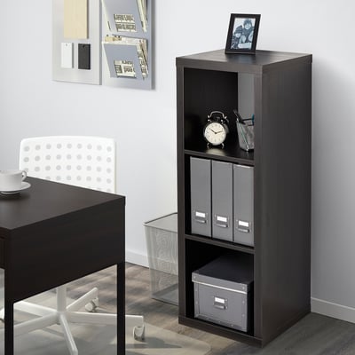 La estantería negra KALLAX se encuentra cerca de un escritorio, sosteniendo un reloj, cajas y una foto. Los estantes son modulares y almacenan los artículos de forma ordenada.