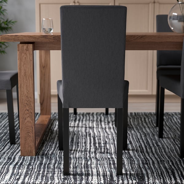 Silla tapizada de color gris oscuro con respaldo alto y patas metálicas empujada debajo de una mesa de madera, adecuada para cenas cómodas.