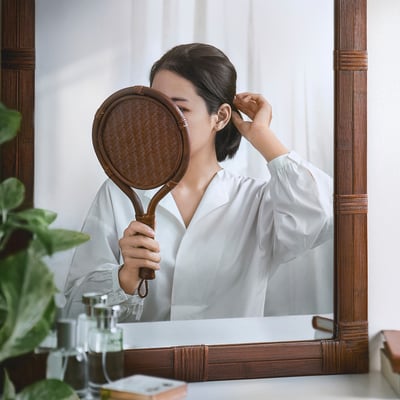 La mujer que sostiene un espejo con mango de ratán revisa la oreja con pinzas, lo que refleja una mezcla de cuidado de belleza y decoración natural.