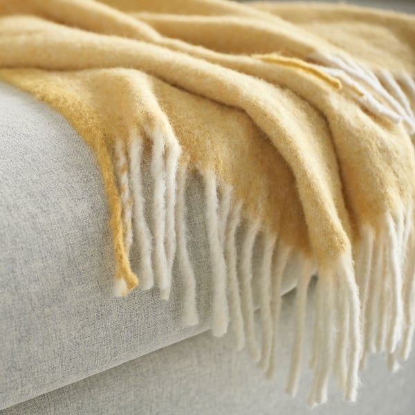 Manta suave de color beige claro con flecos sobre una almohada ligera.