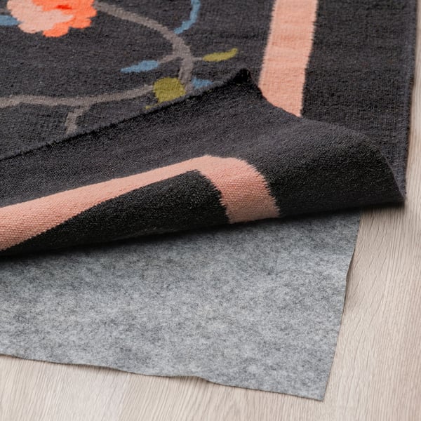 Alfombra de lana tejida a mano con diseño floral sobre gris oscuro, que se muestra plegada con base antideslizante sobre suelo de madera.