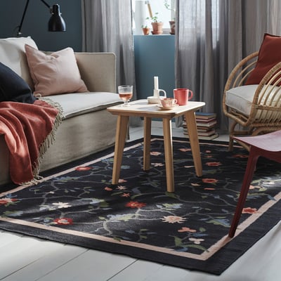 Sala de estar con sofá gris, mesa auxiliar de madera y alfombra negra con diseño floral.