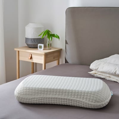 Almohada gris KLUBBSPORRE sobre la cama, más grande que la mesita de noche con lámpara y planta.