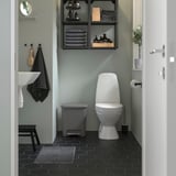 Baño negro con inodoro blanco, papelera gris, paredes verdes, baldosas negras, toallas en silla y estantería.