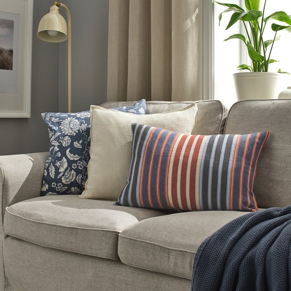 Acogedor sofá de tono neutro con cojines azul oscuro, florales y a rayas. Iluminado por una lámpara de pie y una planta verde junto a la ventana.