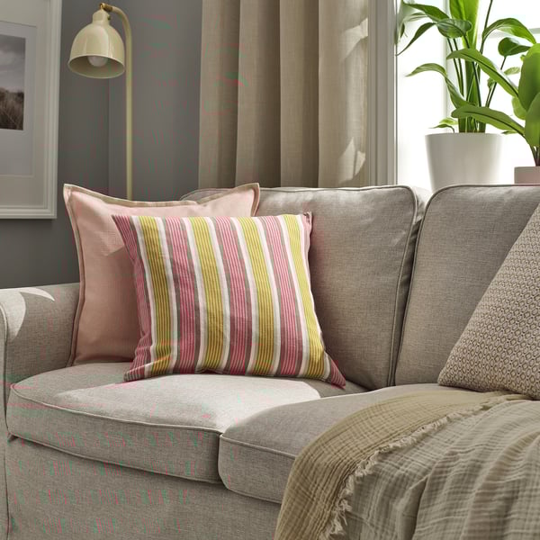 Coloridas almohadas a rayas en una silla acolchada, incluida KORALLBUSKE, con lámpara y planta cerca.