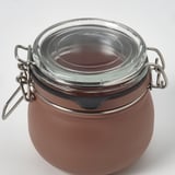 Tarro vacío y hermético con cierre metálico y tapa de cristal. Cuerpo redondo, de color marrón y superficie lisa. Sellado para conservar el contenido.