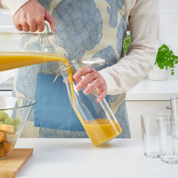 La persona vierte jugo de naranja de una jarra de vidrio etiquetada como KORKEN en vasos transparentes sobre una mesa blanca con frutero.