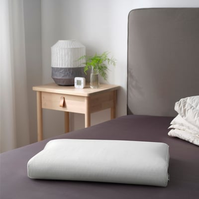 Dormitorio moderno con cabecero gris, mesita de noche de madera, lámpara, reloj, planta verde y almohada ergonómica.