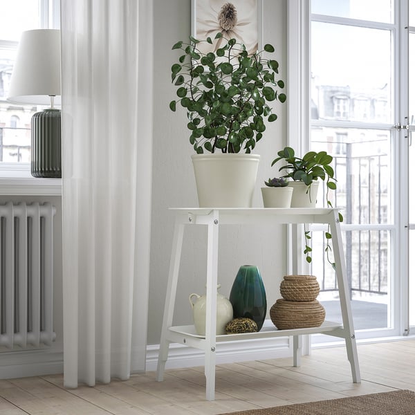 Estante blanco de dos niveles junto a la ventana. Arriba: planta grande, abajo: jarrones, cestas, lámpara, radiador. Minimalista, fresco.