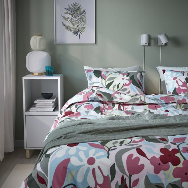 Acogedor dormitorio con ropa de cama floral KORSKOVALL, pared verde, estampado de hojas, lámparas modernas, mesita de noche blanca y cristal turquesa.