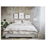 Dormitorio minimalista con ropa de cama blanca, tres cuadros de arte en la pared, una gran planta frondosa a la izquierda y cortinas blancas transparentes a la derecha.