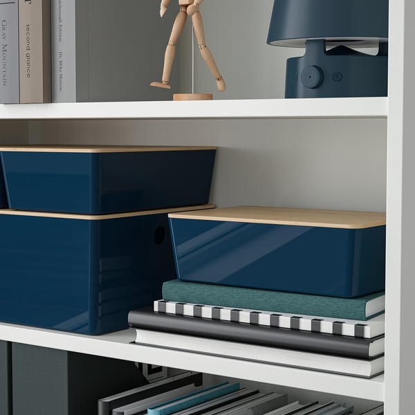 Una estantería blanca con cajas de almacenamiento azules y tapas de madera organiza varios objetos, incluyendo libros, una figura de madera y un altavoz.