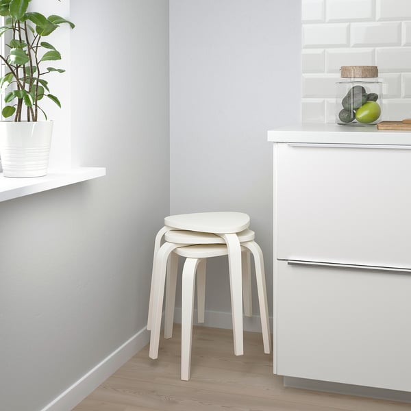 Una cocina moderna con dos taburetes blancos apilados y una planta en maceta. Los taburetes son apilables y tienen un asiento triangular y tres patas para mayor estabilidad.