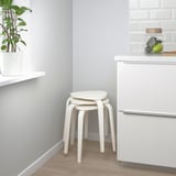 Una cocina moderna con dos taburetes blancos apilados y una planta en maceta. Los taburetes son apilables y tienen un asiento triangular y tres patas para mayor estabilidad.