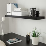 Encuentre un elegante estante de pared bLACK LACK con libros blancos y una cámara vintage, combinado con un escritorio bLACK con un cuaderno apilado y una lámpara moderna.