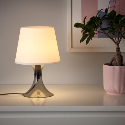 Pequeña lámpara en el mostrador, luz cálida, pantalla blanca, base moderna, crea un ambiente acogedor.