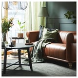 Sala de estar con sofá de cuero marrón, almohadas verdes, lámpara, mesa de centro con velas, flores y alfombra de yute.