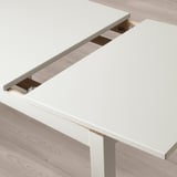 Mesa de comedor extensible LANEBERG blanca con hoja almacenada debajo, con capacidad para 4-6 personas, sobre suelo de madera.