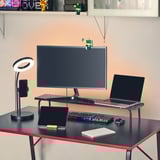 Un soporte para monitor de juegos con teclado, mouse y computadora portátil sobre un escritorio negro. Lámpara led y tablet cerca.