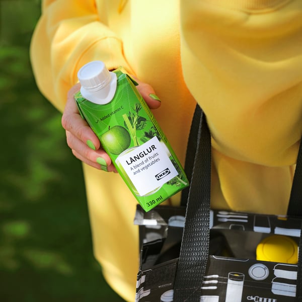 Persona que sostiene un cartón de bebida de batido länglur verde con una gorra blanca, vistiendo una chaqueta amarilla y sosteniendo una bolsa de compras reutilizable en blanco y negro.