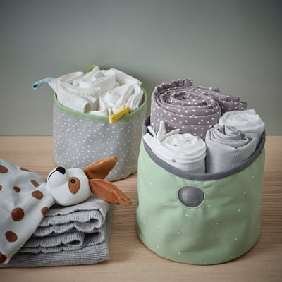 Dos contenedores de almacenamiento de tela, uno verde y otro gris con lunares blancos, que sostienen ropa de bebé enrollada y un animal de peluche sobre una mesa de madera.