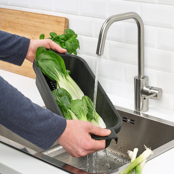 Persona lavando verduras en colador LILLHAVET negro debajo del grifo del fregadero de plata.