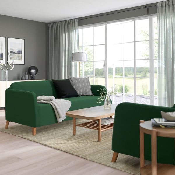 Sala de estar moderna con sofá verde, almohadas, mesa de centro, sillón, mesa auxiliar con libros y alfombra sobre piso de madera clara.