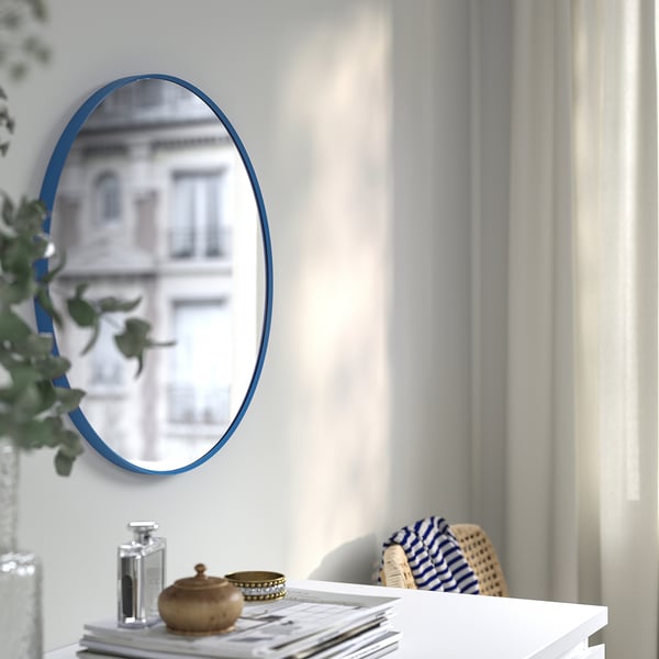 Un espejo ovalado con un marco azul en una pared, que refleja el exterior de un edificio, colocado sobre una mesa blanca con una lámpara y elementos de decoración.