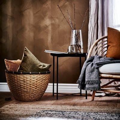 La cesta de bambú tejido LÖVRÄFSA sostiene almohadas cerca de una silla de ratán con una mesa auxiliar negra con un jarrón con ramas; Decoración acogedora y natural.