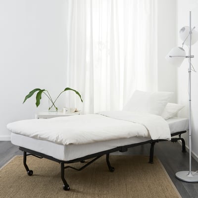Dormitorio minimalista con silla-cama blanca, suelo oscuro, alfombra beige, planta.