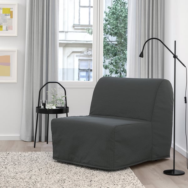 Sillón-cama gris moderno y compacto cerca de la ventana, iluminado por una lámpara de pie alta, junto a un pequeño soporte de plantas redondas.