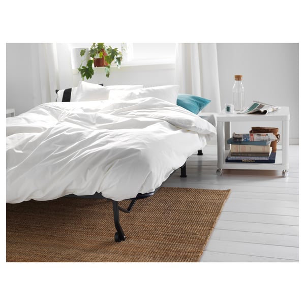 Cama blanca minimalista con patas negras, sábanas frescas, mesita de noche pequeña, libros, botella, planta, suelos blancos, alfombra de yute.