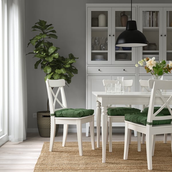 Comedor moderno con mesa de madera blanca, sillas verdes acolchadas, planta grande en una esquina y armario gris.