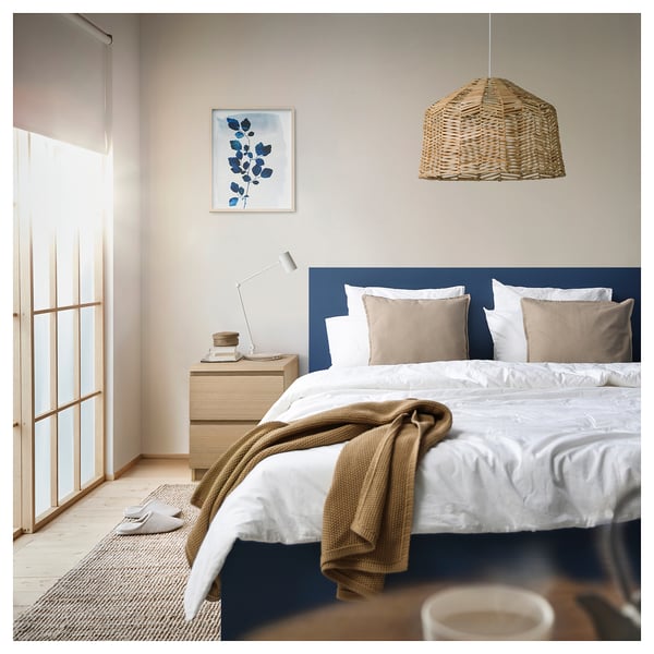 Dormitorio moderno con cama MALM azul, mantas beige, lámpara colgante de ratán y un estampado de pared con follaje azul.