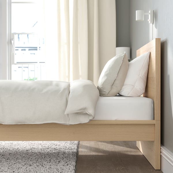 Cama minimalista de roble, cabecero alto, diseño limpio, con ropa de cama y almohadas blancas suaves, junto a un gran ventanal con cortinas ligeras.
