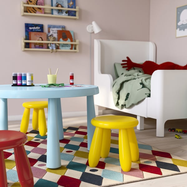 Sala de juegos para niños con mesa azul pastel MAMMUT, pequeños taburetes amarillos y rojos, alfombra colorida, cama blanca con ropa de cama, estantes de pared con libros para niños.