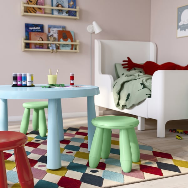 Área de juegos con mesa azul, taburetes verdes, materiales de arte, cama blanca y libros.