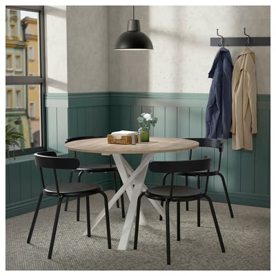 Moderno juego de comedor con mesa redonda, estructura blanca, sillas negras. Acogedor rincón con pared verde y percheros.
