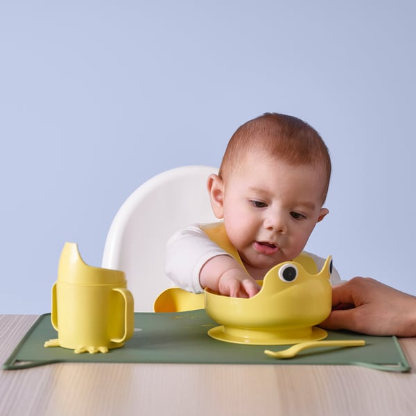 Bebé en silla alta con cuenco de rana. Taza y cuchara a juego sobre tapete verde.