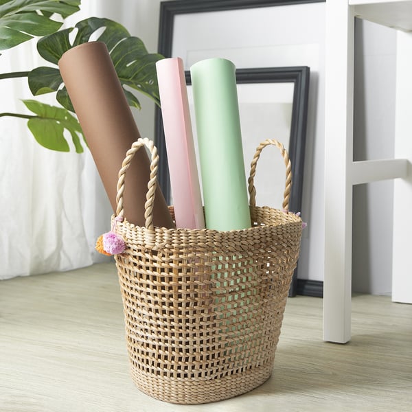 Cesta con telas enrolladas y asas en el suelo junto a la planta, ideal para guardar mantas o cojines.