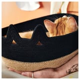 Un gato se asoma de una cesta de yute negro; Textura tejida, forma redonda con recortes triangulares.
