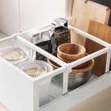 Cajón de cocina organizado con tazones de vidrio, utensilios de madera y rallador de metal en gabinete blanco.