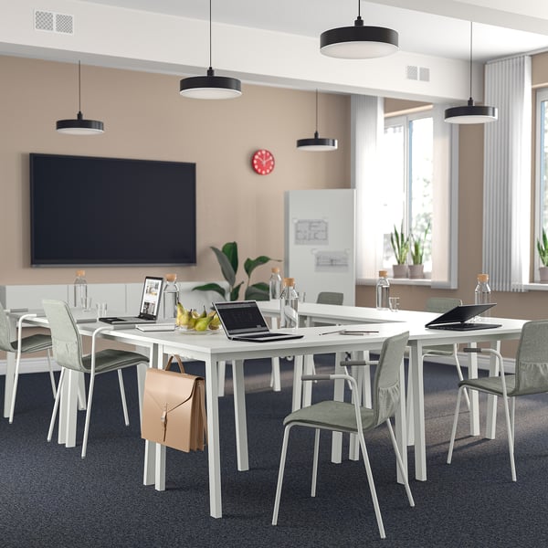 Moderna oficina con mesas blancas, laptops, sillas grises, luces, plantas y pantalla grande.